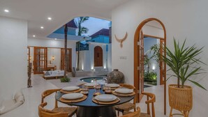 Comedor con una mesa redonda dispuesta para seis personas, rodeada de sillas de mimbre, en un interior moderno con paredes blancas y vistas a un cielo azul y palmeras.