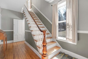 A staircase with wooden steps and white balusters, a wooden table, a door, and a window with curtains. - Broad Street 5 | Androscoggin Main House (Bethel)