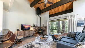 A modern living room with a stone fireplace, a large TV, leather chairs, a sofa, and a balcony with a view of trees. - Cozy lofted condo in Kings Beach close to everything! (Kings Beach)
