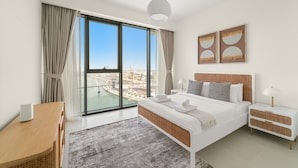 A modern bedroom with a large window, a bed, a bedside table, and a nightstand. - Canal Front Building 9 A - 1001 2BD (Dubai)