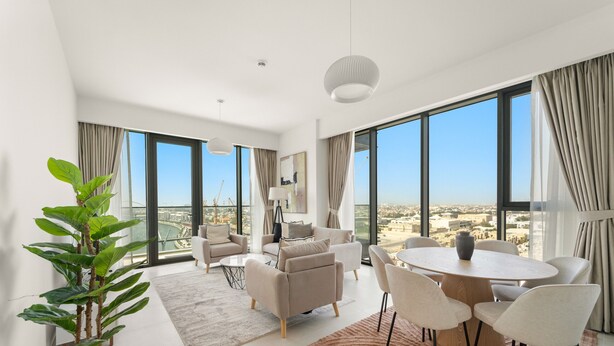 A modern living room with a large window, a potted plant, and a round table with chairs. - Canal Front Building 9 A - 1001 2BD (Dubai)