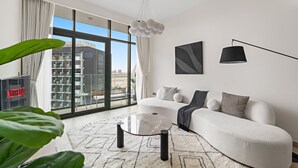 A modern living room with a white sofa, a glass coffee table, and a view of the city through large windows. - Azizi Riviera 3 - 804 1BD (Dubai)
