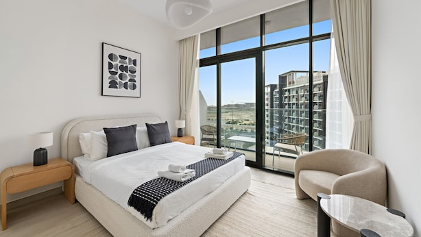 A modern hotel room with a large bed, a side table, a chair, and a view of the city through large windows. - Azizi Riviera 3 - 804 1BD (Dubai)