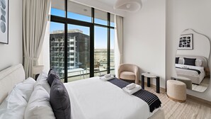 A modern hotel room with a large bed, a sitting area, a balcony with a view of buildings, and a large window. - Azizi Riviera 3 - 804 1BD (Dubai)