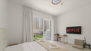 Ein modernes Hotelzimmer mit einem großen Bett, einem Schreibtisch, einem Sessel und Blick auf hohe Gebäude durch das Fenster.