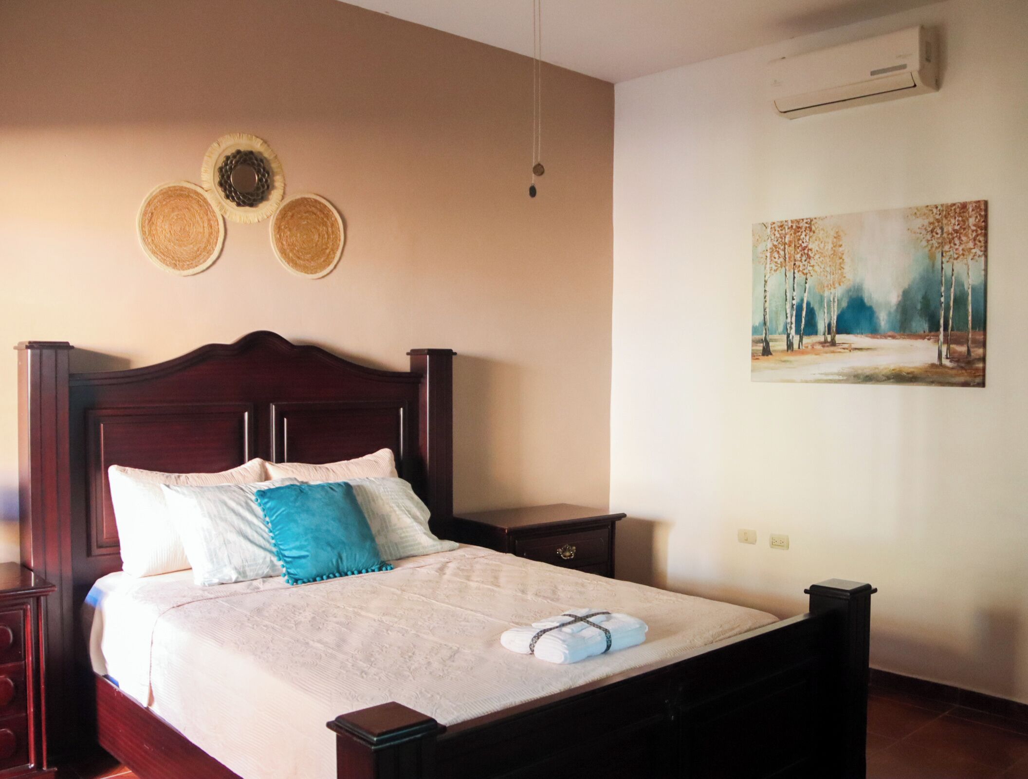 Een netjes opgemaakt bed met een witte dekbedovertrek en blauwe sierkussens, een houten hoofdbord, een nachtkastje met een lamp, en een schilderij van een boslandschap aan de muur.