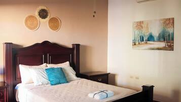 Een netjes opgemaakt bed met een witte dekbedovertrek en blauwe sierkussens, een houten hoofdbord, een nachtkastje met een lamp, en een schilderij van een boslandschap aan de muur.