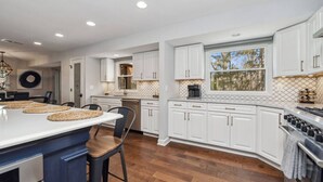 A modern kitchen with white cabinetry, a large island, and a dining area with chairs. - Nautical, Pet-Friendly | Walk to Lake & Downtown (Lake Geneva)