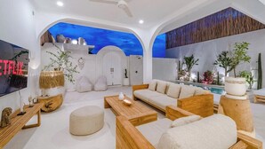 A modern living room with a sofa, coffee table, and a TV. There is a ceiling fan, a fireplace, and a pool area visible.