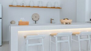 A modern kitchen with a central island, bar stools, a wooden shelf with white objects, a round mirror, and a sink.