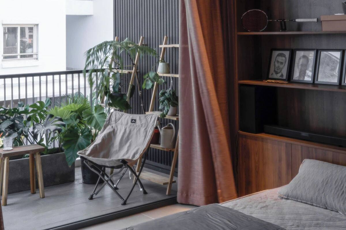 A modern living room with a large window, a balcony with plants, a wooden bookshelf, and a comfortable seating area.
