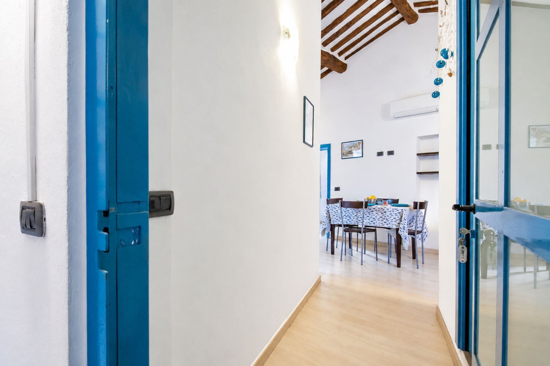 Un pasillo que conduce a un comedor con una mesa dispuesta para cuatro personas, un techo con vigas a la vista y un equipo de aire acondicionado instalado en la pared.