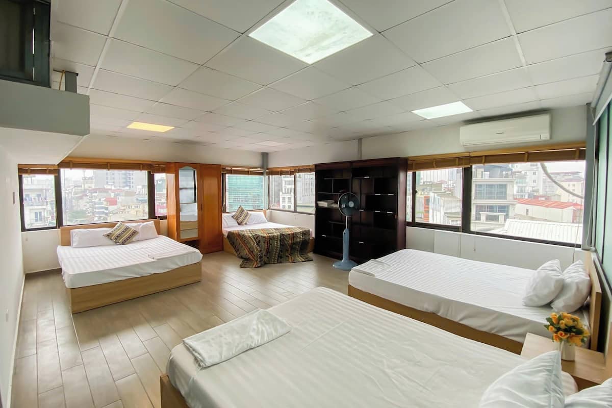 Une chambre d’hôtel avec deux lits, une armoire en bois, un ventilateur sur pied et de grandes fenêtres donnant sur la ville.