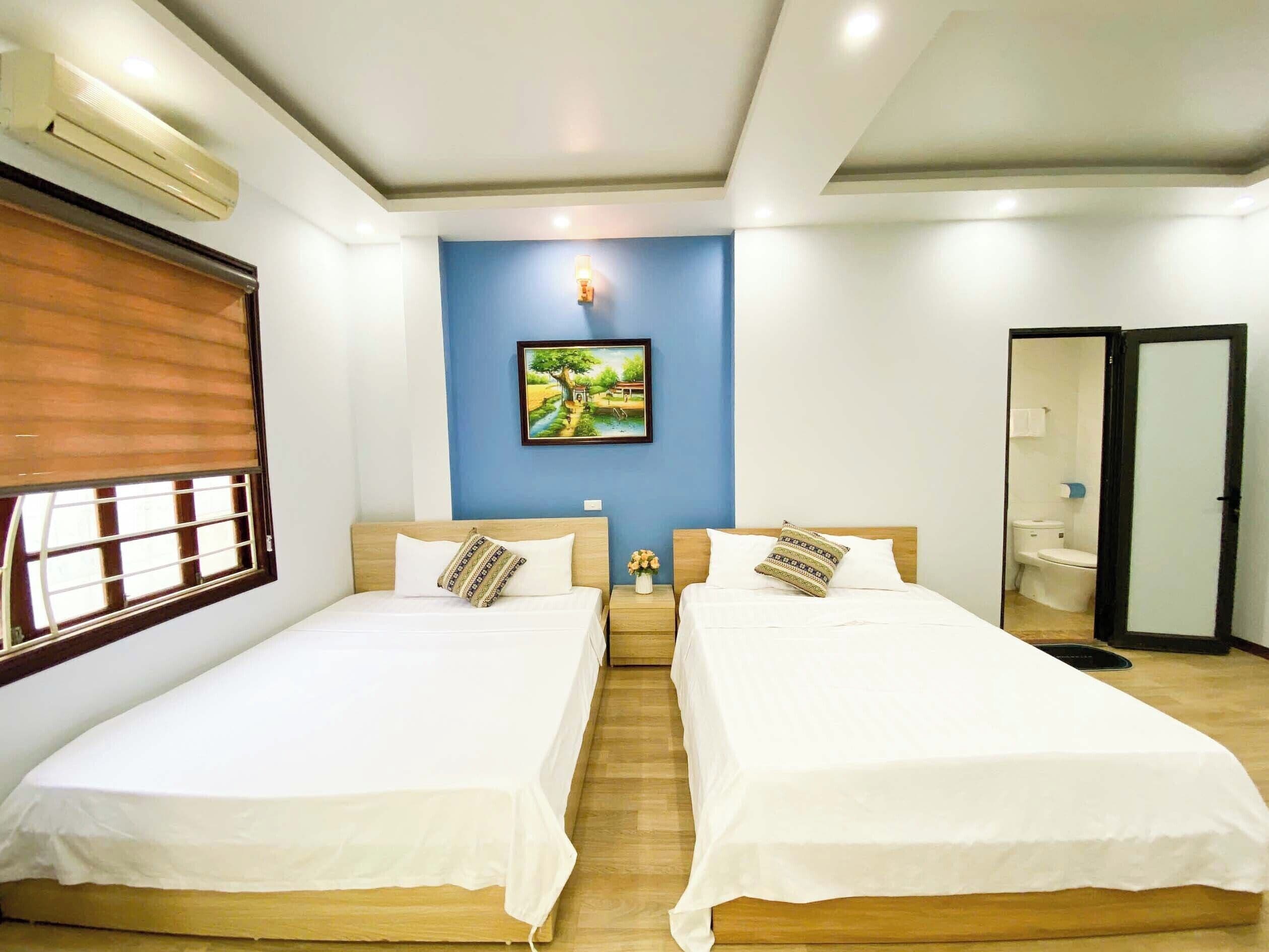 A hotel room with two single beds, a wooden floor, a wall-mounted air conditioner, a painting, and a bathroom visible through an open door.