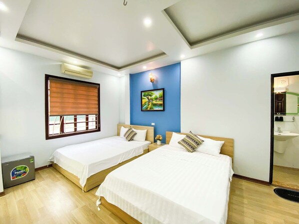 A hotel room with two beds, a window with blinds, a painting on the wall, and a bathroom visible through an open door.