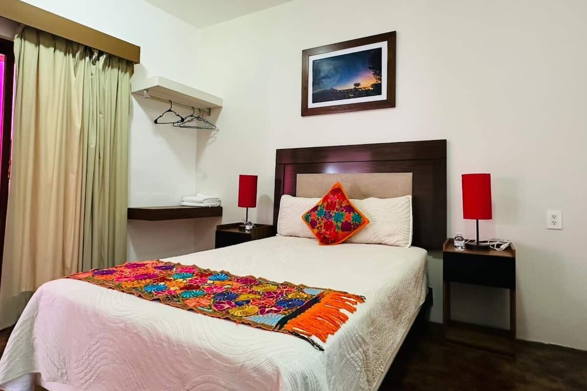 Une chambre d’hôtel avec un grand lit, une table de chevet avec une lampe, une étagère fixée au mur et une fenêtre avec des rideaux.