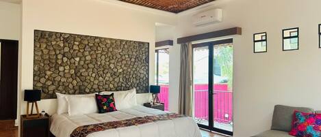 Un dormitorio con pared de piedra, una cama con colcha colorida, un sofá gris y un balcón con valla rosa.