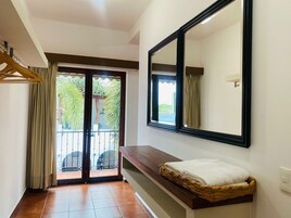 Habitación de hotel con una puerta corrediza de vidrio que da a un balcón, un espejo grande, un banco de madera y una cesta de mimbre con una toalla blanca.