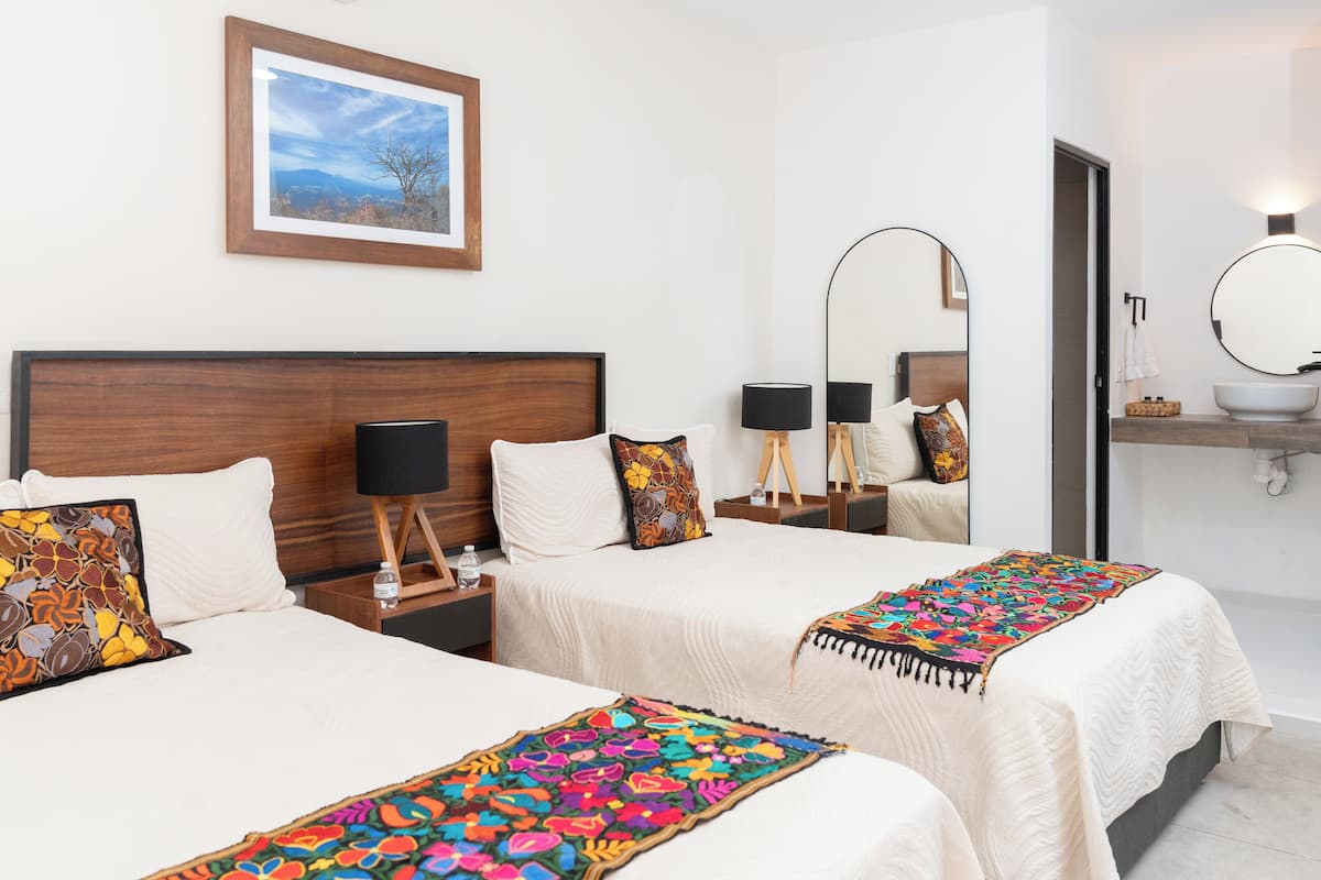 Une chambre d’hôtel avec deux lits, une tête de lit en bois, un tableau encadré au mur, un miroir rond et une table de chevet avec une lampe.