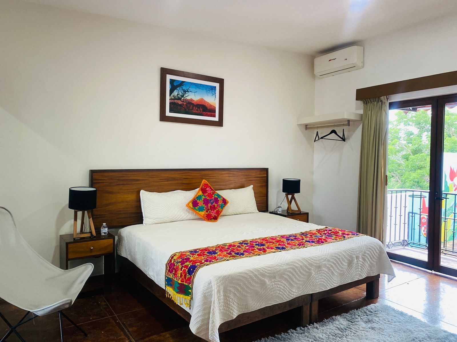 A hotel room with a large bed, a bedside table with a lamp, a chair, a window with a view of greenery, and an air conditioning unit on the wall.