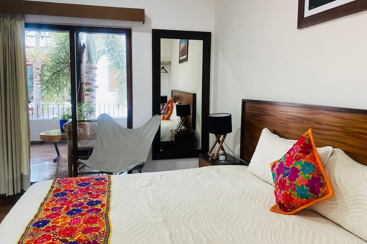 Une chambre d’hôtel comprenant un lit, une chaise, un bureau, un miroir et un balcon avec vue sur les arbres et les bâtiments.