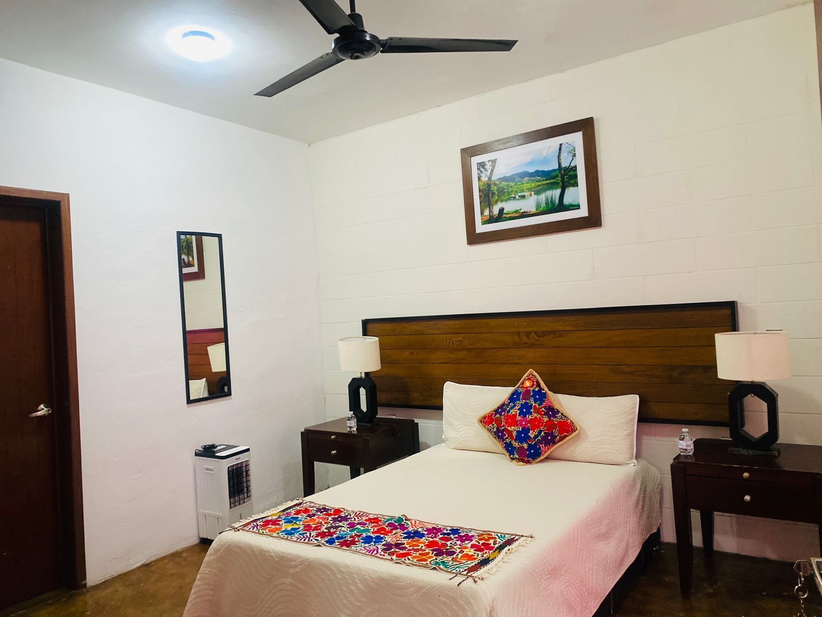 Habitación de hotel con cama, dos mesitas de noche, ventilador de techo, radiador, un cuadro enmarcado en la pared y una puerta.
