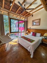 Un dormitorio con cama, una silla, una mesita de noche, un ventilador de techo y una ventana con vistas a edificios.