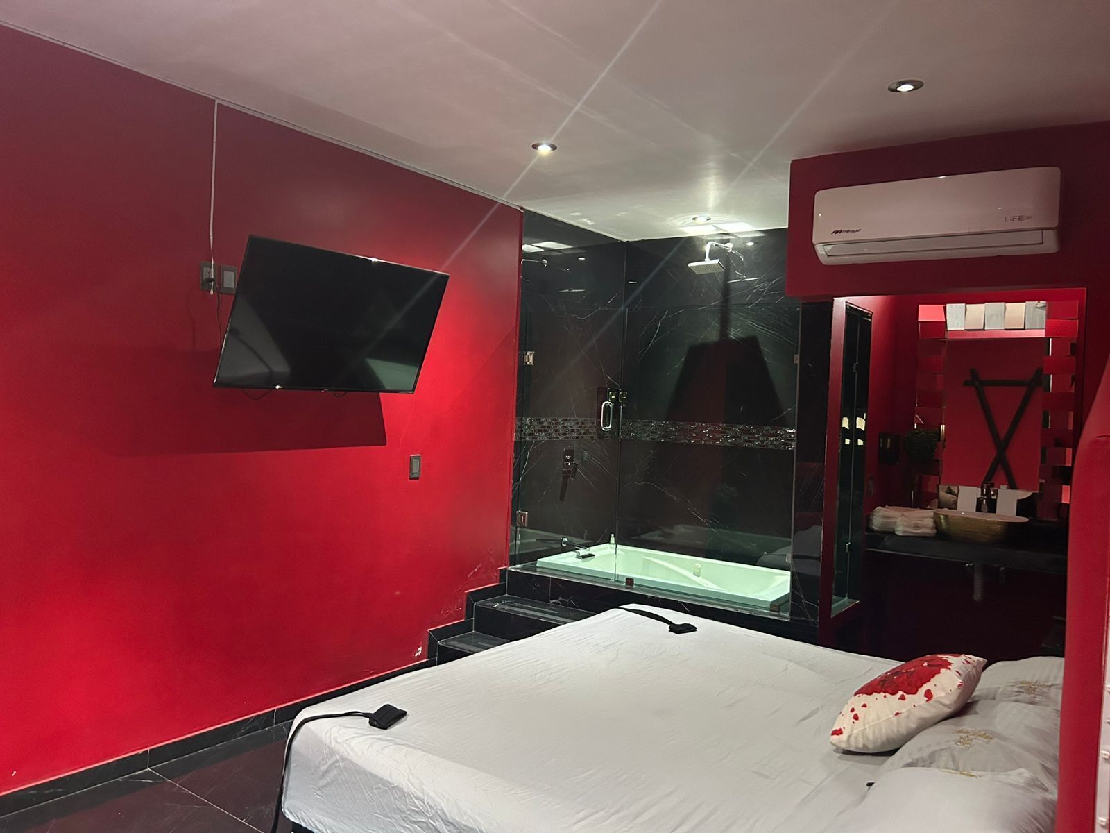 A modern hotel room with a red wall, a large bed, a TV mounted on the wall, a glass-enclosed shower area, and a mirror with a red frame.