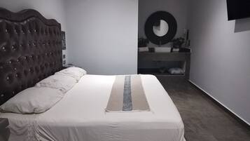 Un dormitorio con cabecera capitonada, cama grande con ropa de cama blanca, espejo redondo en la pared y un banco empotrado con lavabo y espejo.