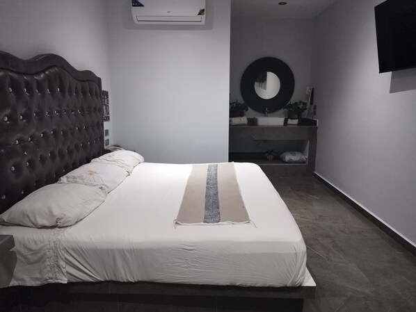 Ein Schlafzimmer mit einer karopoolteren Kopfteil, einem großen Bett mit weißer Bettwäsche, einem runden Spiegel an der Wand und einer eingebauten Sitzbank mit Waschbecken und Spiegel.