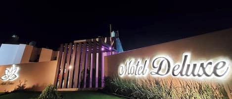 The exterior of the Motel Deluxe with a prominent illuminated sign, a modern design with vertical slats, and a well-maintained lawn.