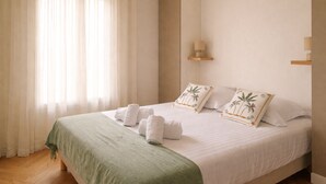 Una cama bien tendida con ropa de cama blanca y detalles verdes, una mesita de noche con lámpara y una ventana con cortinas de gasa.