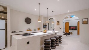 A modern kitchen with a central island, a sink, and a built-in oven. There are stools around the island and a large arched doorway leading to a living area with a TV displaying 'NETFLIX'. - 3BR Rooftop Summary: (Kabupaten Badung)