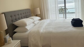 A neatly made bed with white linens, a grey upholstered headboard, and a bedside table with a lamp and a tissue box. - Yallumbee, Seal Rocks - Modern, Private Beach House In Prime Location (Seal Rocks)