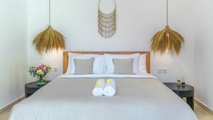 Un lit bien fait, avec du linge de lit blanc et deux serviettes, une table de chevet ornée d’un vase de fleurs et un objet décoratif fixé au mur.