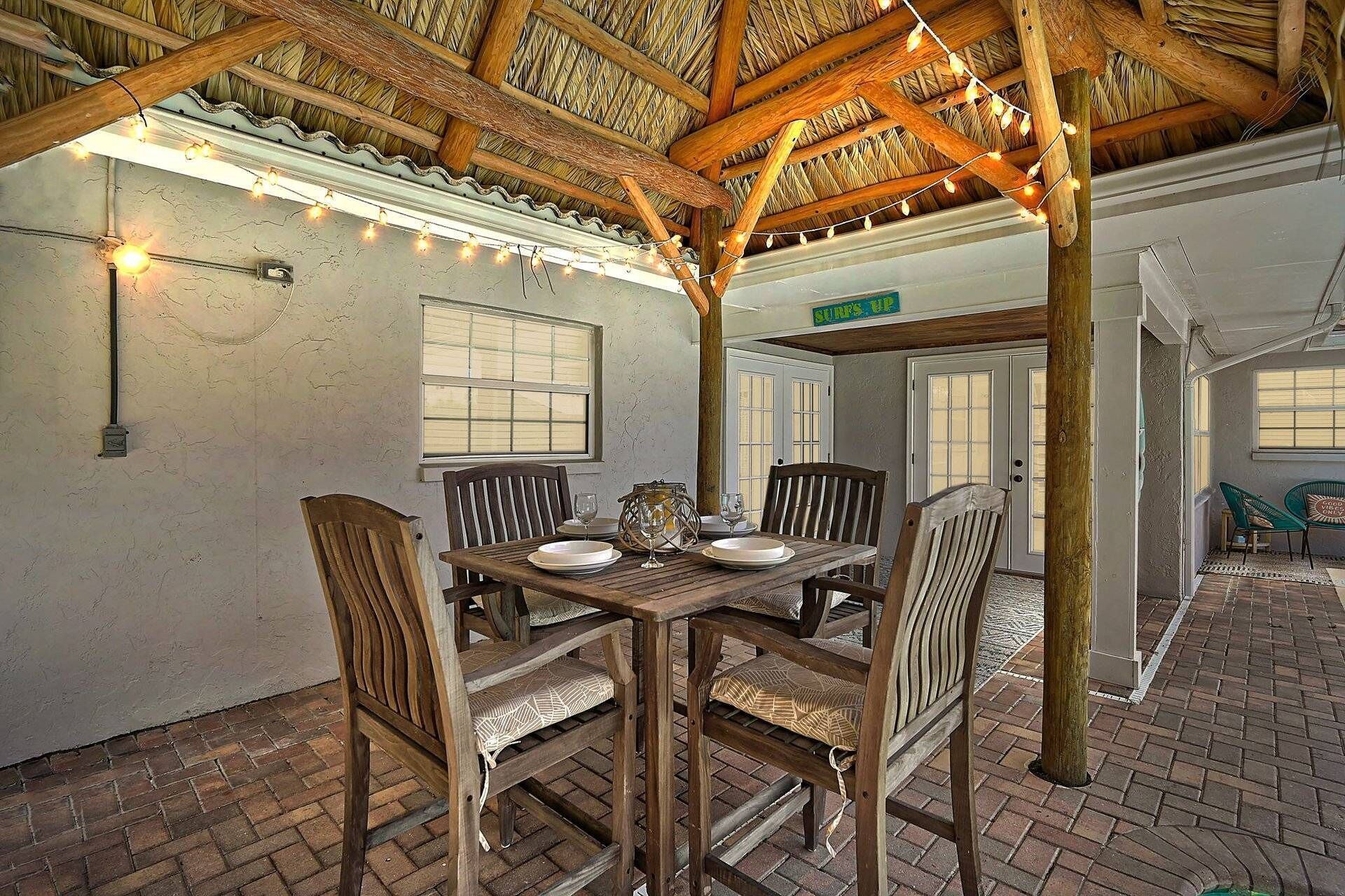 An outdoor dining area with a wooden table and chairs, string lights, and a thatched roof structure.