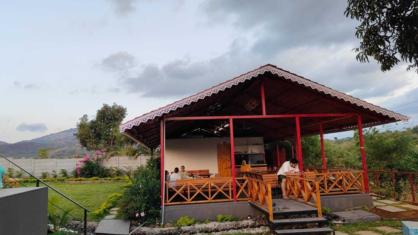 A covered outdoor seating area with wooden benches and a red roof, surrounded by greenery and a mountainous backdrop.