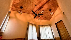 A bedroom with a wooden ceiling, a bed with a patterned bedspread, and two windows with curtains.