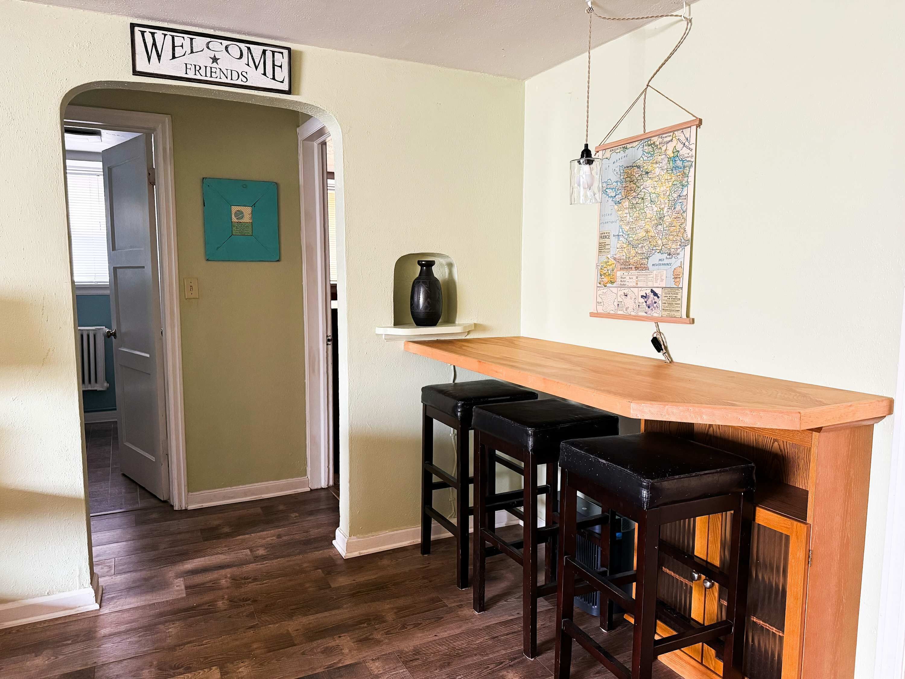 A kitchen with a wooden bar and stools, a 'WELCOME FRIENDS' sign, a map on the wall, and a door leading to another room.