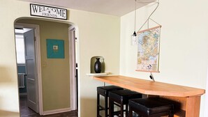 Une cuisine avec un bar en bois et des tabourets, une pancarte indiquant « BIENVENUE, AMIS », une carte accrochée au mur et une porte donnant sur une autre pièce.