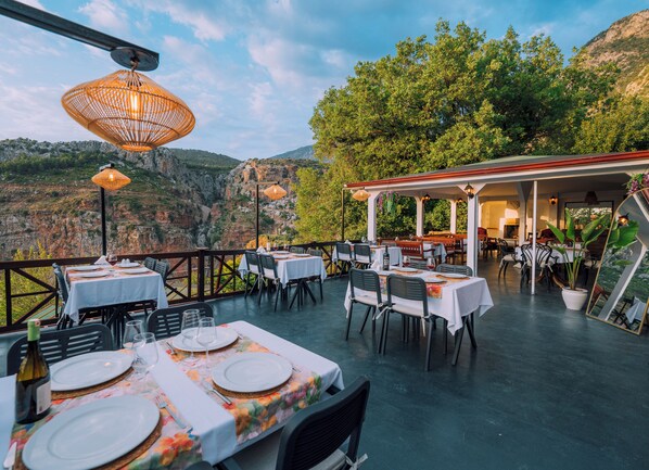 An outdoor dining area with tables set for two, surrounded by chairs, and overlooking a mountainous landscape.