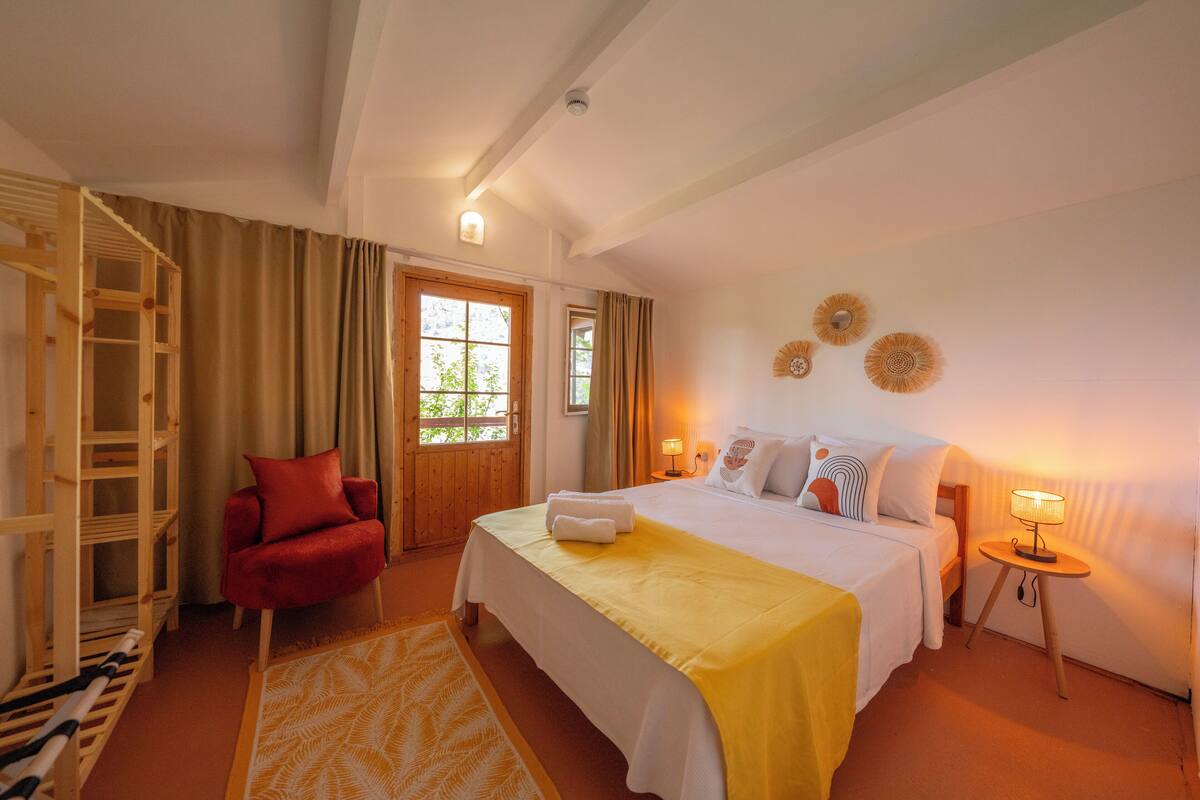 Une chambre d’hôtel avec un lit, une chaise rouge, une échelle en bois, une petite table avec une lampe et une fenêtre avec des rideaux.
