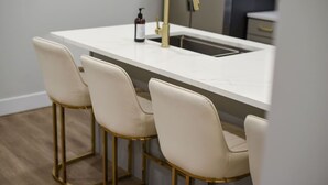 A kitchen with a white countertop, a gold faucet, and three white bar stools with gold legs.