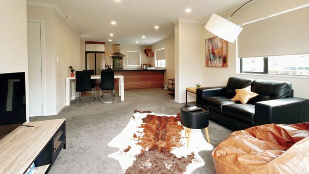 A modern living room with a leather sofa, a dining area with chairs, a kitchen in the background, and a large window. - Canvas of Comfort — A Modern Townhouse of Four Suites (Hamilton)