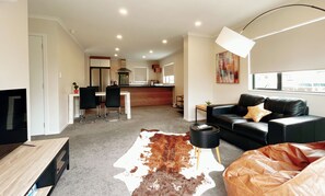 A modern living room with a leather sofa, a dining area with chairs, a kitchen in the background, and a large window. - Canvas of Comfort — A Modern Townhouse of Four Suites (Hamilton)