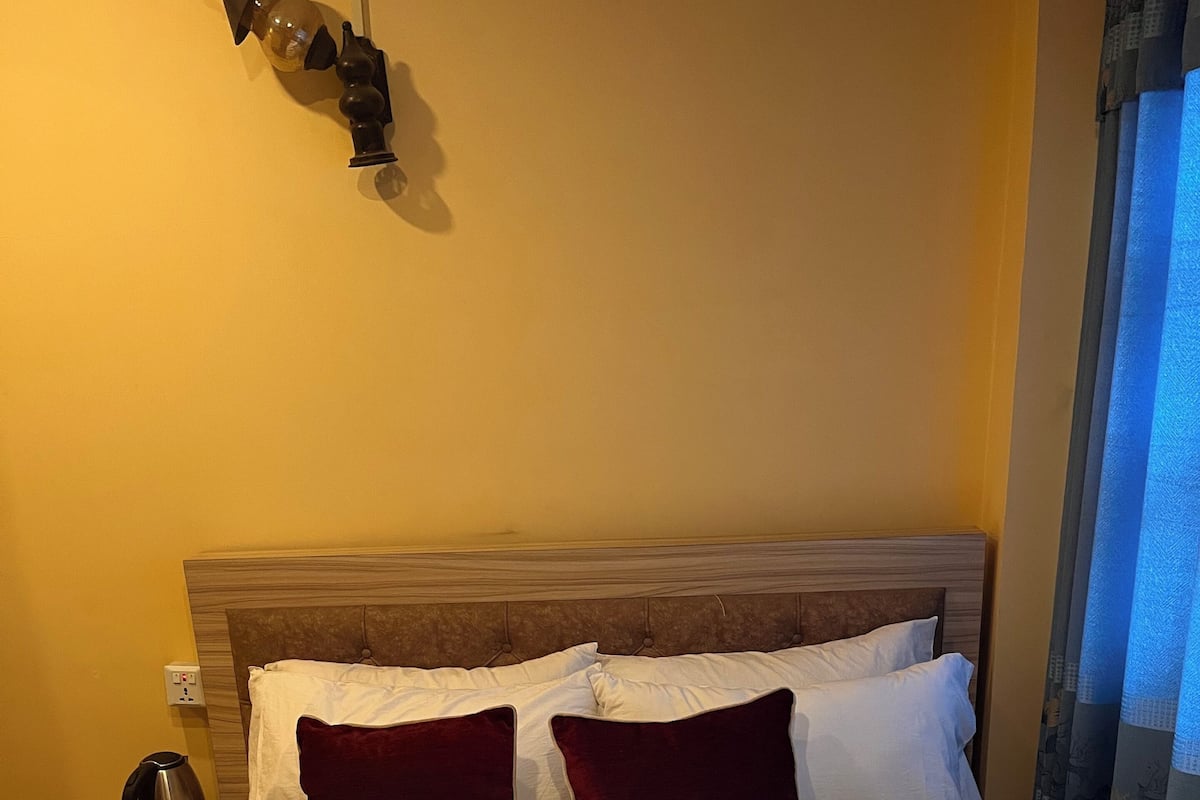Una habitación de hotel con una cama, almohadas, una mesita de noche, una lámpara y un enchufe.