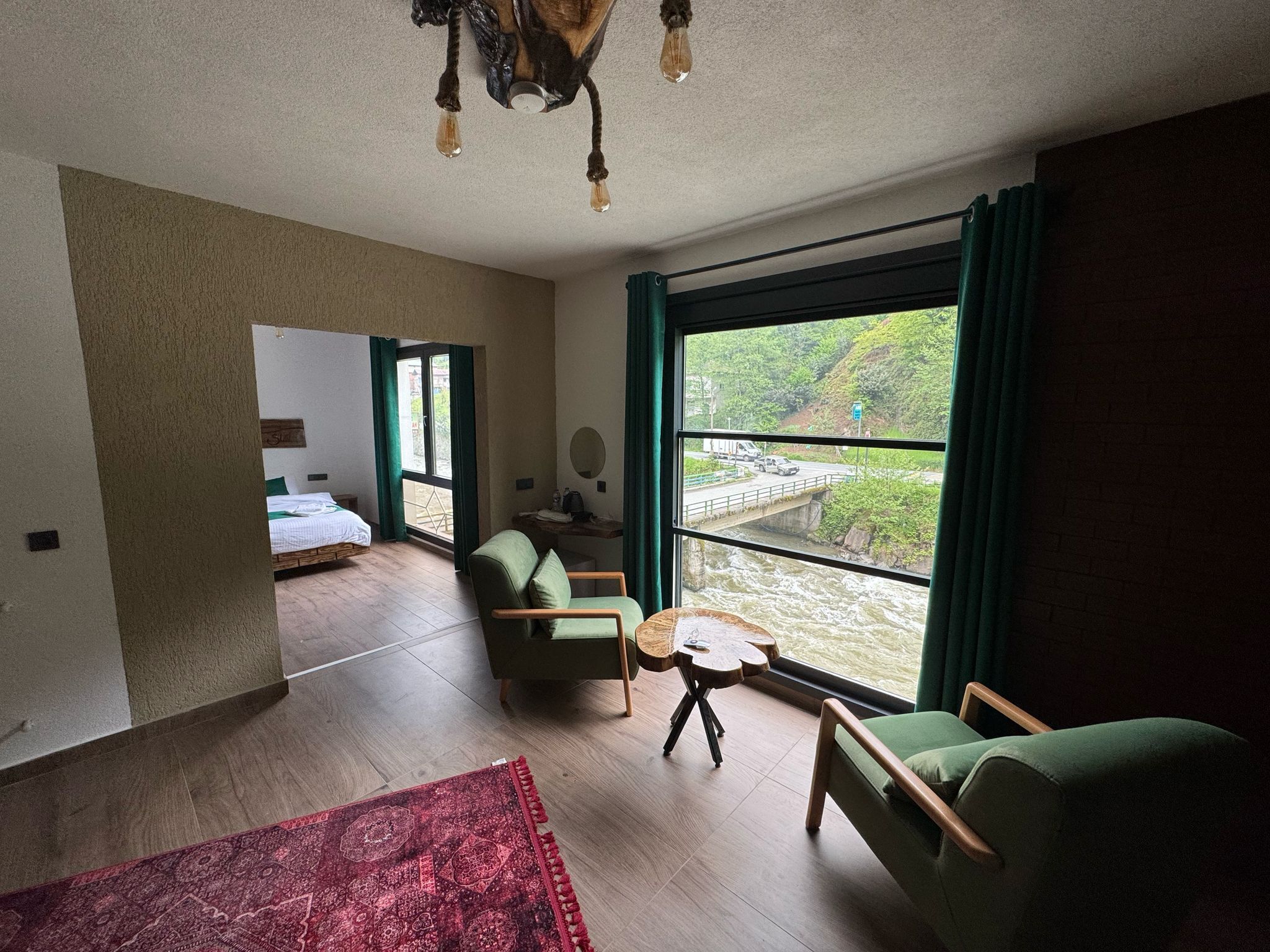 A spacious room with a large window, a bed, a green armchair, a small round table, and a red patterned rug.