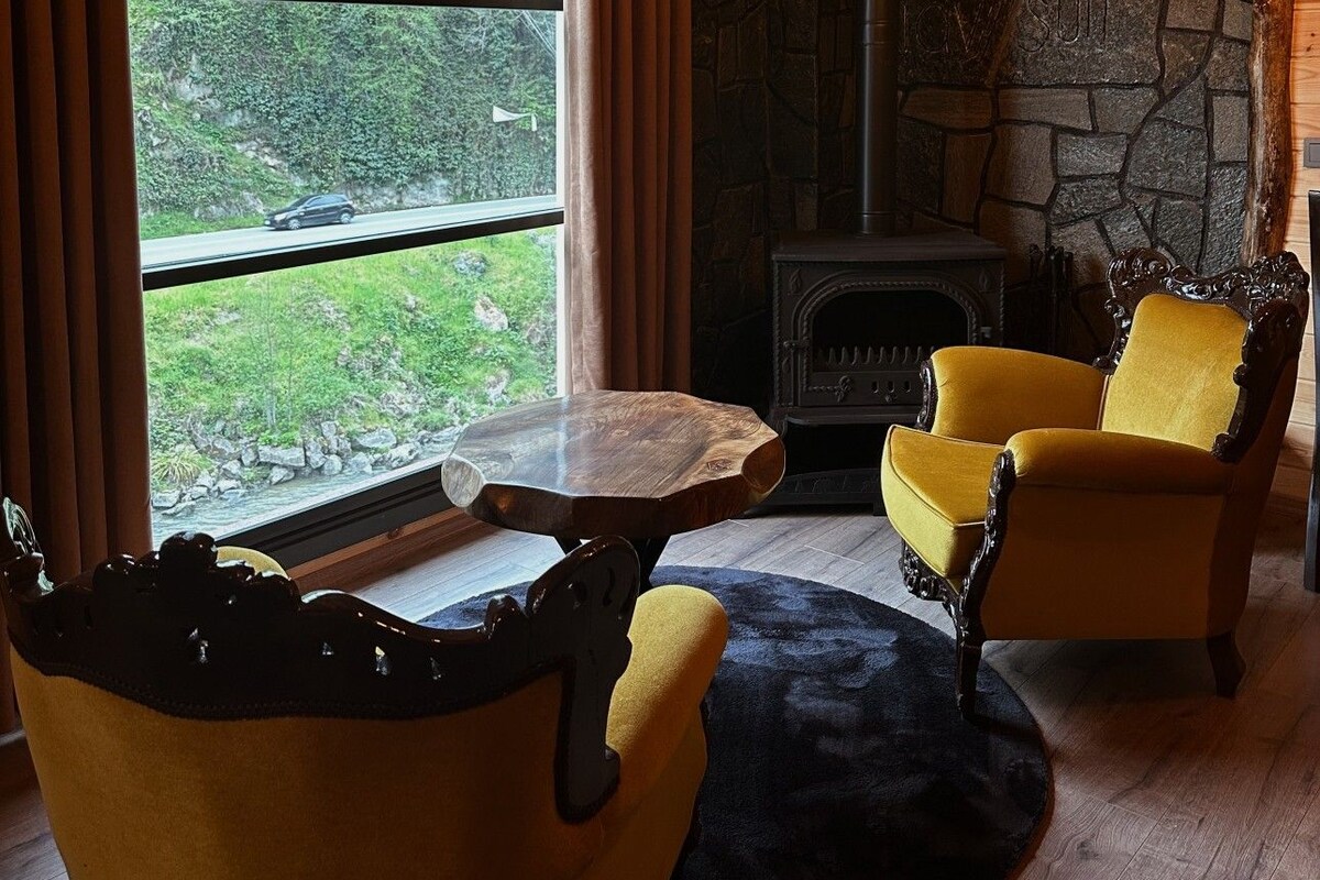 A cozy room with a stone fireplace, wooden ceiling beams, and a large window offering a view of greenery.