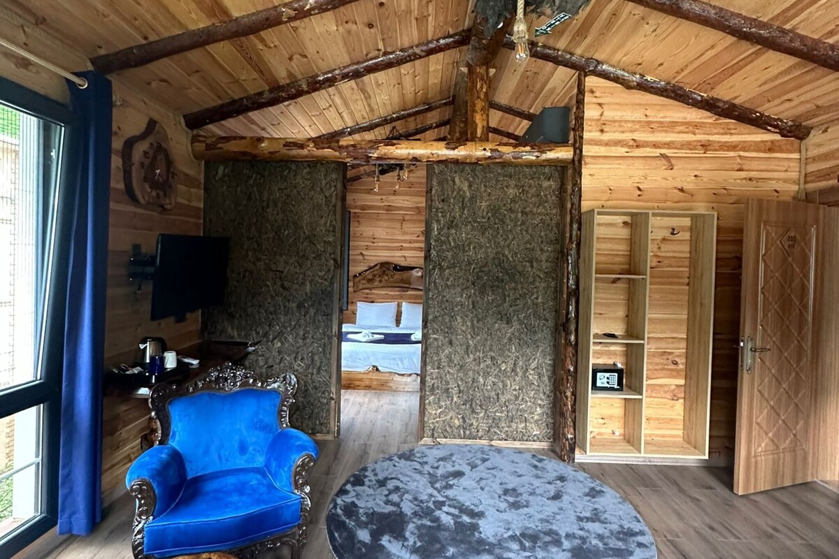 A rustic wooden interior with a round table, blue upholstered chairs, and a bed visible through an open door.