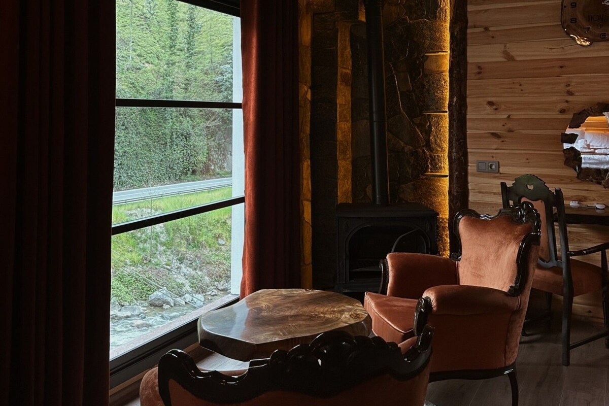 A rustic interior with a stone fireplace, wooden ceiling beams, and a view of greenery outside.